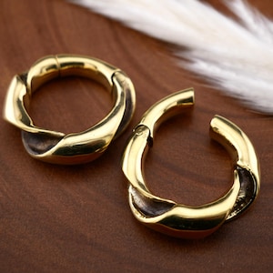 May include: Two gold-colored, twisted hoop earrings. The earrings have a hinged clasp and a dark brown accent on the inner part of the twist. The earrings are displayed on a brown surface.