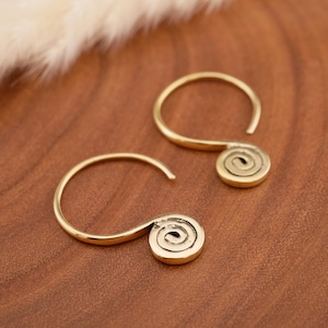 Könnte beinhalten: Ein Paar goldfarbene Creolen mit einem glatten, kreisförmigen Drahtdesign, das an der Basis in einen flachen, spiralförmigen Anhänger übergeht. Die Ohrringe haben eine polierte metallische Oberfläche und eine einfache, offene Hakenstruktur.