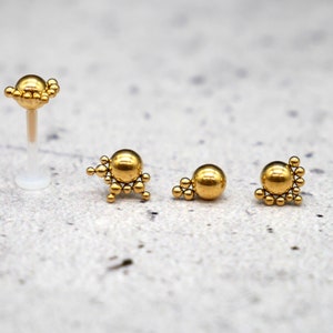 Peut inclure: Trois bijoux de corps dorés avec un groupe de petites boules. Les pièces sont disposées en rangée du plus grand au plus petit.