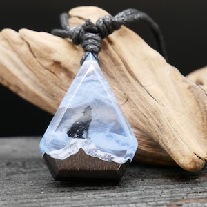 Könnte beinhalten: Eine geometrische Anhänger-Halskette mit einer Wolfssilhouette, die den Mond anheult, eingebettet in klarem Harz mit einem blau-weißen Wolkeneffekt. Der Anhänger hat einen dunklen Holzsockel und hängt an einer schwarzen Kordel.