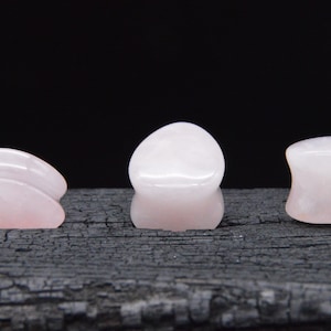 Puede incluir: Tres medidores de oreja de piedra rosa de diferentes formas y tamaños. Los medidores están hechos de cuarzo rosa y son lisos y pulidos.