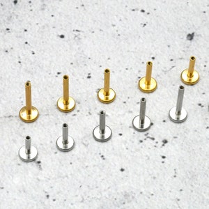 May include: A collection of 12 gold and silver body jewellery pieces. The jewellery is in the shape of a small stud with a flat back. The studs are arranged in two rows on a white surface.