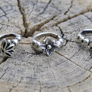 Peut inclure: Trois anneaux de nez en argent avec des motifs décoratifs différents. La première bague présente une fleur de lotus, la deuxième bague a un motif floral et la troisième bague a un symbole stylisé.