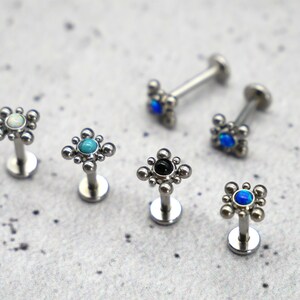 May include: Five silver body jewelry pieces with different colored gemstones. Each piece has a small square design with a round gemstone in the center. The gemstones are white, turquoise, black, blue, and blue.