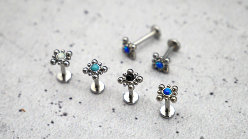 May include: Five silver body jewelry pieces with different colored gemstones. Each piece has a small square design with a round gemstone in the center. The gemstones are white, turquoise, black, blue, and blue.
