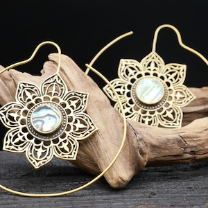 Può includere: Un paio di orecchini a cerchio color oro. Ogni orecchino presenta un design a forma di fiore con intagli intricati e un intarsio centrale in conchiglia di abalone. Gli orecchini sono esposti su un pezzo di legno alla deriva su sfondo nero.