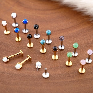 Può includere: Una collezione di piercing per labbra con pietre preziose colorate, tra cui bianco, blu, nero, turchese, viola e verde. I piercing hanno perni dorati o argentati e sono esposti su una superficie di legno.