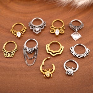 Può includere: Una collezione di piercing per il naso e septum in tonalità oro e argento. Gli anelli presentano vari design, tra cui filigrana, pietre preziose e dettagli a catena. I gioielli sono esposti su una superficie marrone.