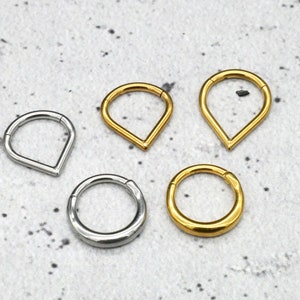 Peut inclure: Un assortiment de bijoux de corps, dont deux anneaux clicker dorés et deux argentés, ainsi qu'un anneau articulé en forme de goutte doré. Les bijoux sont présentés sur une surface grise clair mouchetée.
