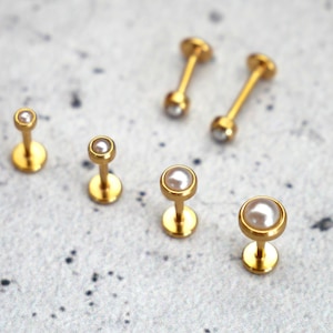 Könnte beinhalten: Eine Sammlung goldfarbener Labret-Ohrstecker mit Perlen- und Kristallakzenten. Der Schmuck wird auf einer gesprenkelten grauen Oberfläche präsentiert. Die Ohrstecker variieren in Größe und Design und zeigen eine minimalistische Ästhetik.