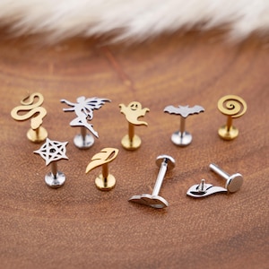 May include: A selection of lip and ear studs in various designs. The studs feature a snake, fairy, ghost, bat, spiral, spiderweb, and leaf. The studs are made of silver and gold-coloured metal.