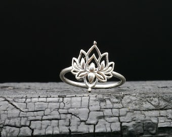 Puede incluir: Un anillo de plata con un delicado diseño de flor de loto. El anillo está sobre una superficie oscura y texturizada.