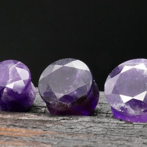 Może przedstawiać: Trzy zatyczki do uszu z ametystu, każda o fasetowanym, zaokrąglonym kształcie. Zatyczki są ciemnofioletowe i ułożone w rzędzie na ciemnej, fakturowanej powierzchni.