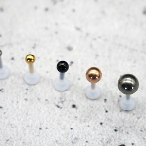 Könnte beinhalten: Ein Set mit sechs Labret-Steckern mit unterschiedlichfarbigen Kugeln. Die Kugeln sind silber, gold, schwarz, roségold und gunmetal.