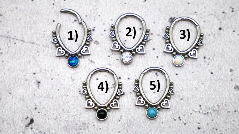 Puede incluir: Cinco anillos de septum de plata con un dise&ntilde;o de coraz&oacute;n y piedras preciosas de diferentes colores. Los anillos est&aacute;n numerados del 1 al 5.