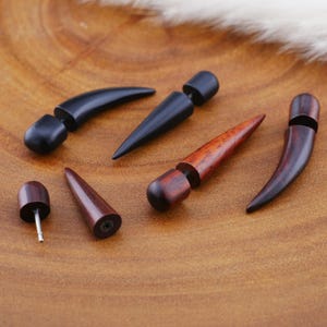 Può includere: Sei dilatatori per orecchie in legno di varie forme e colori. I dilatatori sono realizzati in legno scuro e legno chiaro. Le forme includono una forma curva, una forma appuntita e una forma arrotondata.