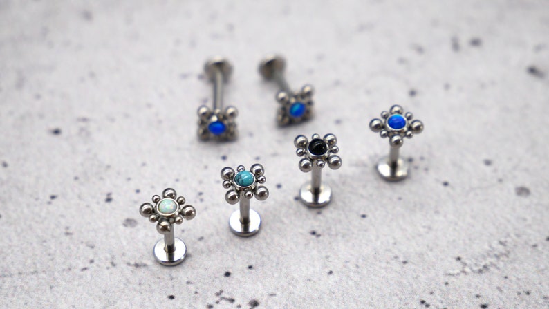 May include: Six silver body jewelry pieces with a square design and a single gemstone in the center. The gemstones are blue, turquoise, black, and white.