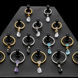 Könnte beinhalten: Eine Auswahl von 12 goldenen, silbernen und schwarzen Captive-Bead-Ringen mit baumelnden Kristall-Anhängern. Die Anhänger sind in verschiedenen Farben, darunter klar, blau, lila und schwarz.