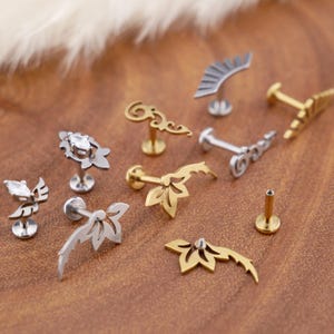 Op de afbeelding: Een collectie metalen lichaamsieraden in zilver- en goudtinten, met diverse decoratieve vormen zoals bladmotieven, vleugelontwerpen, filigraan krullen en waaiers, sommige geaccentueerd met kleine heldere marquise-geslepen edelstenen.