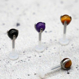 Könnte beinhalten: Vier verschiedene farbige Edelstein-Körper-Schmuckstücke. Der Schmuck ist aus klarem Acryl und Edelstahl gefertigt. Die Edelsteine sind schwarz, lila, braun und orange.