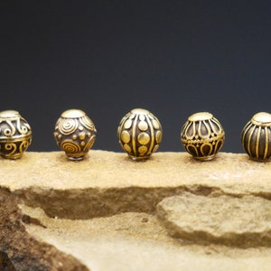 Può includere: Cinque perline di metallo color oro con motivi intricati su una superficie di roccia marrone.