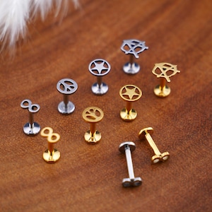 Puede incluir: Una colección de joyas para el cuerpo de plata y oro con varios diseños, incluyendo símbolos de infinito, estrellas, signos de paz y huellas de patas. Las joyas se muestran sobre una superficie de madera.