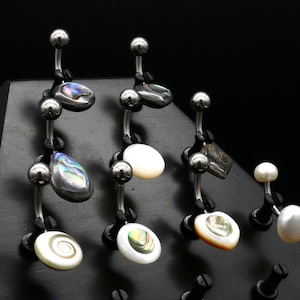 May include: Assortment of belly button rings with various iridescent and white shell and pearl accents. The rings are displayed on a black triangular stand.