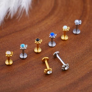 Könnte beinhalten: Sortiment von acht Körperschmuckstücken aus Silber und Gold mit kleinen blütenförmigen Anhängern und bunten Edelsteinen. Jedes Stück hat einen kleinen Stift und eine Kugel am Ende.