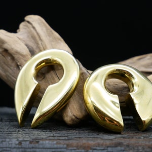 May include: Two gold-colored metal ear gauges with a unique teardrop shape and a small opening in the center. The gauges are on a wooden surface.