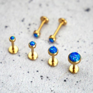 Könnte beinhalten: Fünf goldene Körperschmuckstücke mit blauen Opal-Steinen. Der Schmuck ist in einer Reihe auf einer hellgrauen Oberfläche angeordnet.