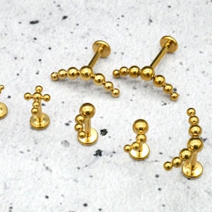 Könnte beinhalten: Ein Set mit sechs goldfarbenen Körperschmuckstücken. Der Schmuck verfügt über eine Vielzahl von Designs, darunter eine Ansammlung kleiner Kugeln, eine Kreuzform und eine geschwungene Linie aus kleinen Kugeln. Der Schmuck ist aus Chirurgenstahl gefertigt und für verschiedene Piercings geeignet.