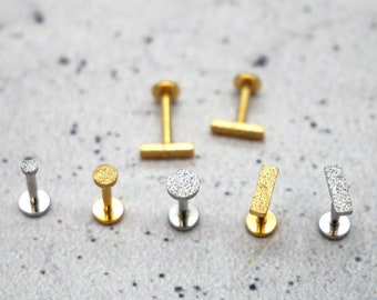 Puede incluir: Un conjunto de siete piezas de joyería para el cuerpo, que incluye dos tachuelas de barra doradas, dos tachuelas redondas doradas, una tachuela redonda plateada y dos tachuelas de barra plateadas. Todas las piezas tienen un acabado brillante y texturizado.