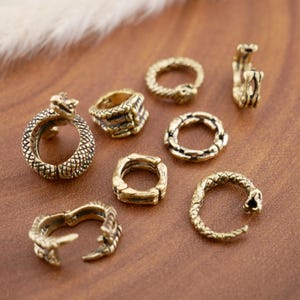 Puede incluir: Una colección de anillos dorados antiguos con diseños de serpientes. Los anillos varían en estilo, algunos con una forma de serpiente completa, otros con texturas y formas de serpiente. Los anillos se exhiben sobre una superficie marrón.
