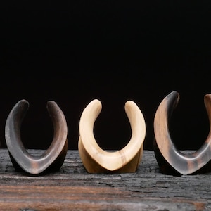 May include: A set of six wooden ear gauges in various sizes, with a curved, horseshoe-like shape. The gauges are made of light and dark wood, creating a natural, earthy aesthetic.