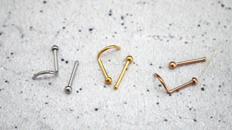 May include: Three nose rings with different styles and colors. One is silver with a curved design, one is gold with a curved design, and one is rose gold with a straight design.