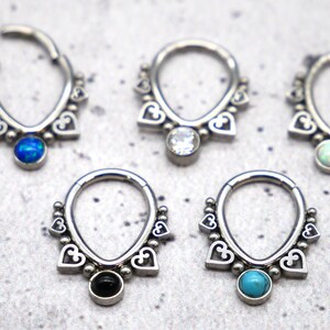 Puede incluir: Cinco anillos de septum plateados con un dise&ntilde;o de coraz&oacute;n y piedras preciosas de diferentes colores. Las piedras preciosas son azules, transparentes, blancas, negras y turquesa.
