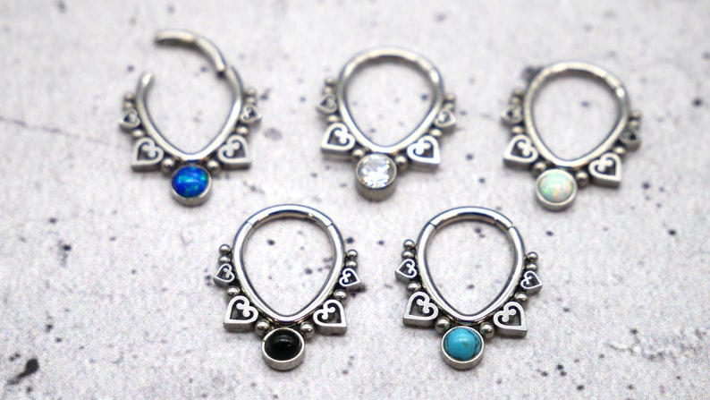 Puede incluir: Cinco anillos de septum plateados con un dise&ntilde;o de coraz&oacute;n y piedras preciosas de diferentes colores. Las piedras preciosas son azules, transparentes, blancas, negras y turquesa.