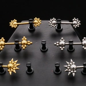 May include: Six pairs of nipple rings displayed on a black triangular stand. The rings are made of stainless steel and gold, with different designs, including starburst and geometric patterns.