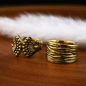 Könnte beinhalten: Zwei goldfarbene Ringe. Ein Ring hat ein strukturiertes, wellenförmiges Design. Der andere Ring hat ein geometrisches Design mit einem erhabenen Muster aus Kreisen und Linien.