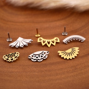 Könnte beinhalten: Eine Sammlung von sechs dekorativen Ohrstecker-Aufsätzen in Gold- und Silbertönen, mit verschiedenen Fächer-, Bogen- und geometrischen Ausschnittdesigns. Drei Metallstifte mit flachen Rückseiten sind hinter den Schmuckstücken platziert.