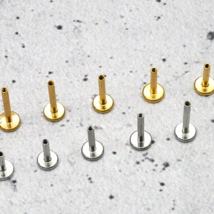 May include: A collection of 14 gold and silver body jewellery pieces. The jewellery is in the shape of a small stud with a flat base. The studs are arranged in two rows on a light grey surface.