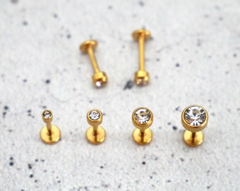 Puede incluir: Un conjunto de seis piezas de joyería corporal de color dorado con acentos de cristal transparente. La joyería incluye una variedad de tamaños y estilos, incluyendo tachuelas y pesas.
