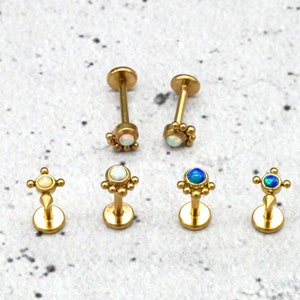 Op de afbeelding: Zes goudkleurige lichaamsieraden met opaal edelstenen. Twee stuks hebben een enkele opaal edelsteen met een halo van kleine kralen. Twee stuks hebben een enkele opaal edelsteen met een kruis van kleine kralen. Twee stuks hebben een enkele opaal edelsteen met een cirkel van kleine kralen.