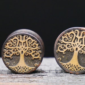 May include: Three wooden ear gauges with a tree of life design. The gauges are different sizes and are made of dark wood with a light brown tree design.
