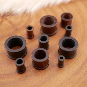 Könnte beinhalten: Eine Sammlung von neun dunkelbraunen Ohr-Plugs aus Holz in verschiedenen Größen. Jeder Plug hat eine zylindrische Form mit einem hohlen Zentrum und ausgestellten Rändern an beiden Enden. Das Holz weist eine glatte, polierte Oberfläche auf.