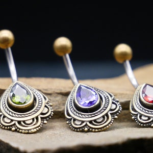 May include: Three belly button rings with ornate gold settings and teardrop-shaped gemstones. The gemstones are green, purple, and red.