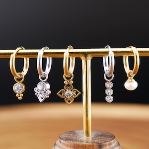 May include: A display of six earrings, each with a unique design. The earrings are gold and silver-toned, featuring various charms like a teardrop, a floral design, and a pearl. The earrings are displayed on a gold-colored stand.