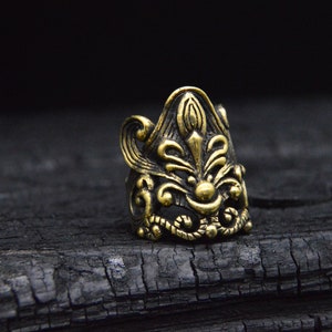 Könnte beinhalten: Ein goldfarbener Ring mit einem komplizierten, verzierten Design, das ein stilisiertes Blumenmotiv zeigt. Der Ring ist vor einem dunklen, strukturierten Hintergrund platziert.