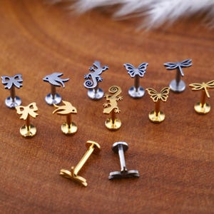 Könnte beinhalten: Ein Set mit 11 Körperschmuckstücken aus Silber und Gold mit verschiedenen Designs, darunter Schmetterlinge, Libellen, Eidechsen und Schleifen. Der Schmuck wird auf einer braunen Holzoberfläche präsentiert.