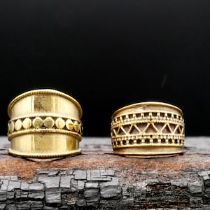 Op de afbeelding: Twee gouden ringen met ingewikkelde ontwerpen. De ring aan de linkerkant heeft een band met kleine cirkels, terwijl de ring aan de rechterkant een geometrisch patroon van driehoeken heeft.